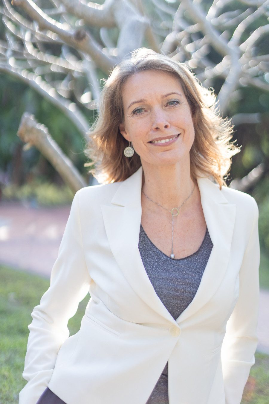 Woman in white jacket smiling outdoors with trees behind.