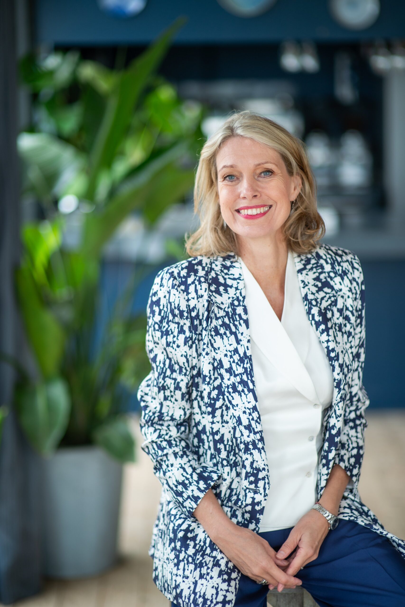 Smiling woman in a white and blue blazer near greenery.