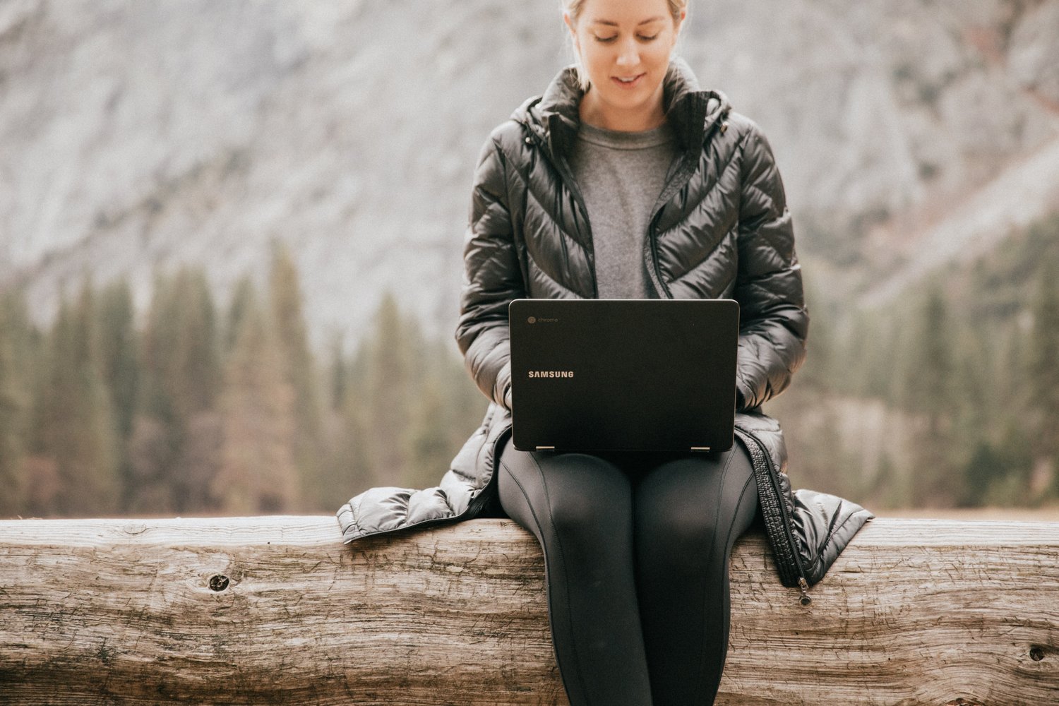 Woman in a black coat using a laptop on a log in nature.