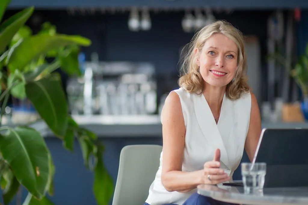 Smiling woman in a white top sitting at a table indoors.